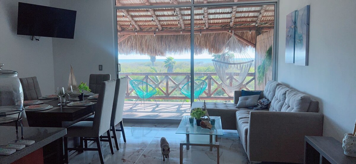 A well-appointed living area features large glass doors leading to a spacious balcony with a thatched roof. A dining table and comfortable seating are present, complemented by a relaxed atmosphere created by hanging hammocks visible outside.