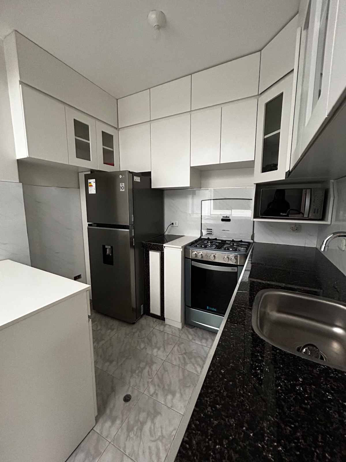 A compact kitchen features sleek white cabinetry and a dark granite countertop. Stainless steel appliances, including a refrigerator and a gas stove, are prominently displayed. A single sink is situated under the window, with cabinetry overhead providing additional storage.