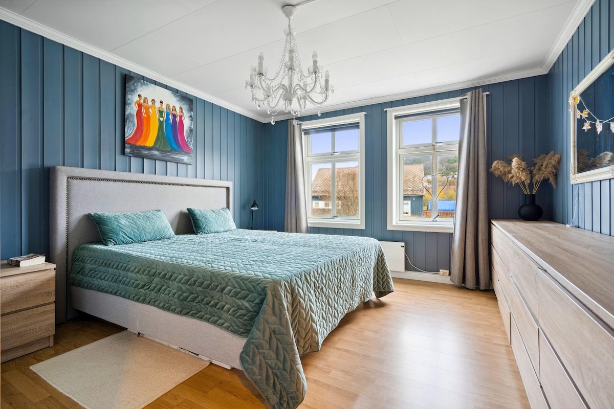 A well-appointed bedroom features deep blue wall paneling and a spacious bed with a light teal coverlet. Natural light enters through two windows, and a colorful wall art piece adds a pop of color. A chandelier hangs overhead, complementing the wooden furnishings.
