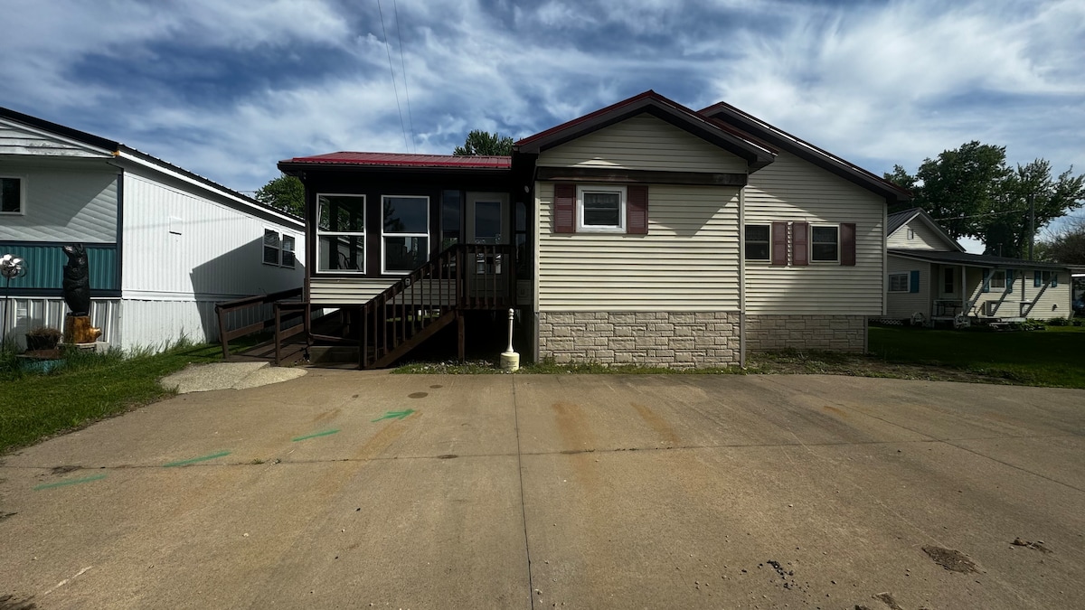 The exterior of the cottage features a front porch with a ramp and steps leading to the entry. A wide driveway allows parking for several vehicles, with a grassy area on one side and neighboring homes visible in the background. The sky is partially cloudy.