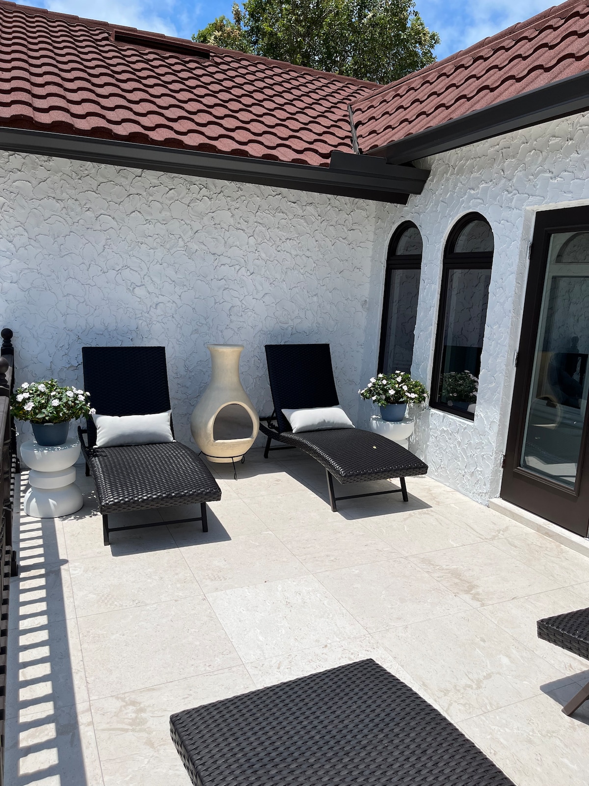 An outdoor patio space features two lounge chairs with plush cushions. Potted plants on either side provide greenery, and a decorative ceramic piece stands nearby. The wall is finished in textured white stucco, complementing the terracotta roof above under a clear blue sky.