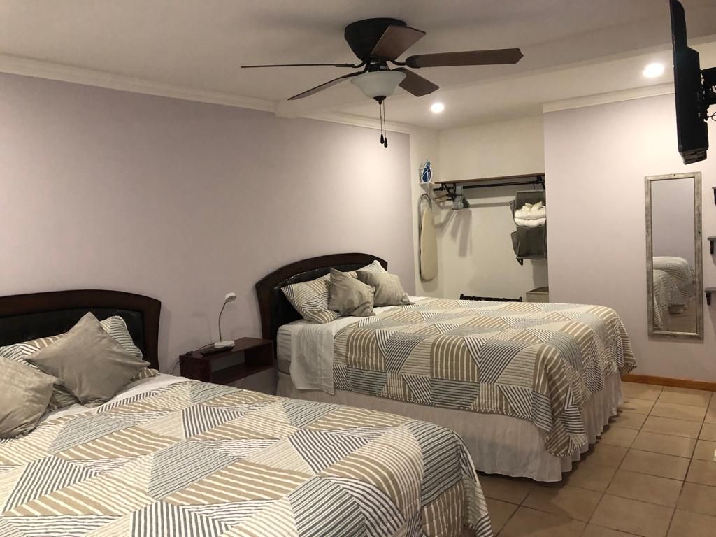 A spacious room features two neatly made beds with patterned bedding, complemented by side tables with lamps. A ceiling fan hangs above for comfort. A large mirror reflects the room, while a coat rack and neatly stacked towels are visible in the background.
