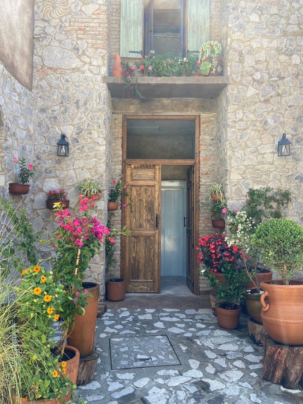 An inviting entrance is framed by natural stone walls and adorned with vibrant flowerpots. Lush plants and colorful blooms create a welcoming atmosphere, leading to a sturdy wooden door. Soft lighting is provided by hanging lanterns, enhancing the charm of the entryway.