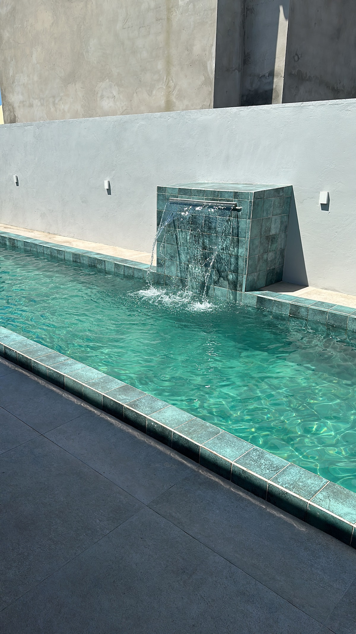 A semi-Olympic swimming pool is viewed, featuring a tranquil waterfall that flows from a stone wall. The clear turquoise water is complemented by a tiled border and surrounding smooth, dark flooring, creating a serene and refreshing environment.