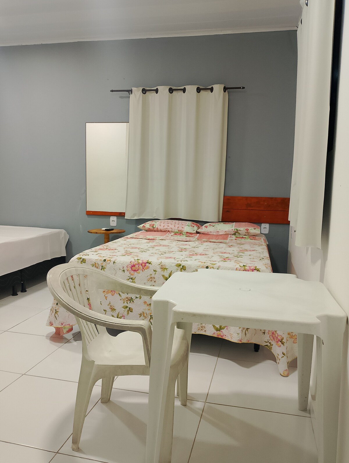 A guest room features a floral-patterned bedspread covering a spacious bed, accompanied by a simple white chair and table. A mirror hangs above the table, and light filters in through a window dressed with light curtains, while a contrasting dark wall adds depth to the space.