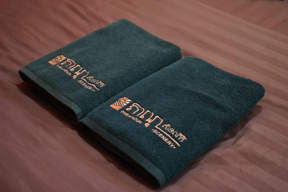 Two neatly folded dark teal towels, featuring an embroidered gold logo, are placed on a striped bedspread. The towels are soft and inviting, emphasizing the thoughtful details provided for guests.
