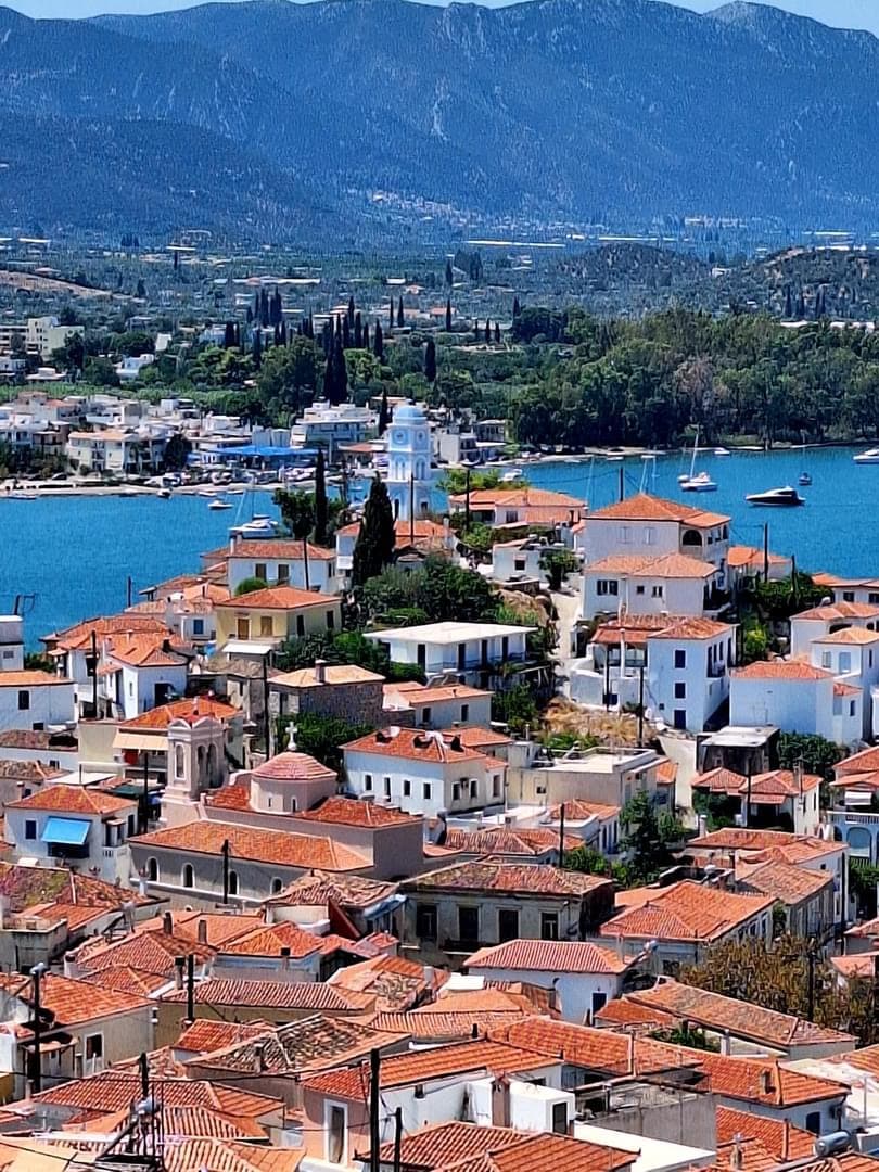 A panoramic view reveals a picturesque coastal town characterized by white and terracotta buildings nestled along a turquoise bay. Prominent features include the tall blue-domed church and surrounding green hills, with boats dotting the water enhancing the tranquil scene.