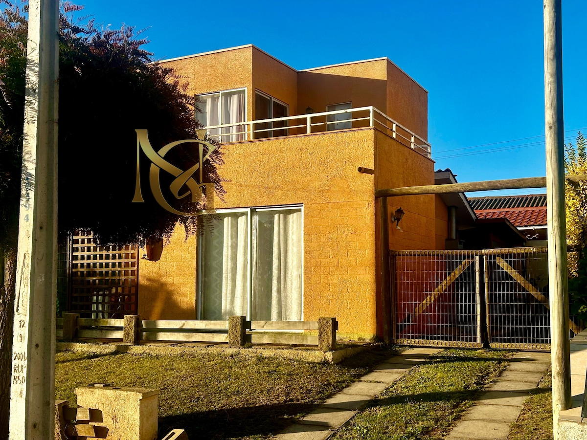 A two-story yellow house is shown, featuring large windows that allow natural light to fill the space. A balcony extends from the upper floor, providing outdoor access. A gate and a pathway are visible, leading to the entrance, which is framed by a lush tree.