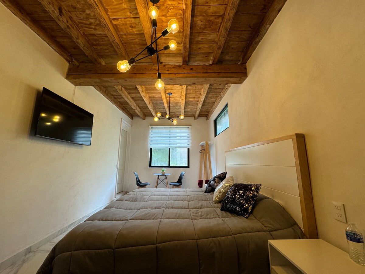 A spacious bedroom features wooden ceiling beams and modern lighting. A large bed with a neutral-toned coverlet is the focal point, complemented by decorative pillows. Natural light enters through windows, illuminating a seating area with two chairs and a small table.