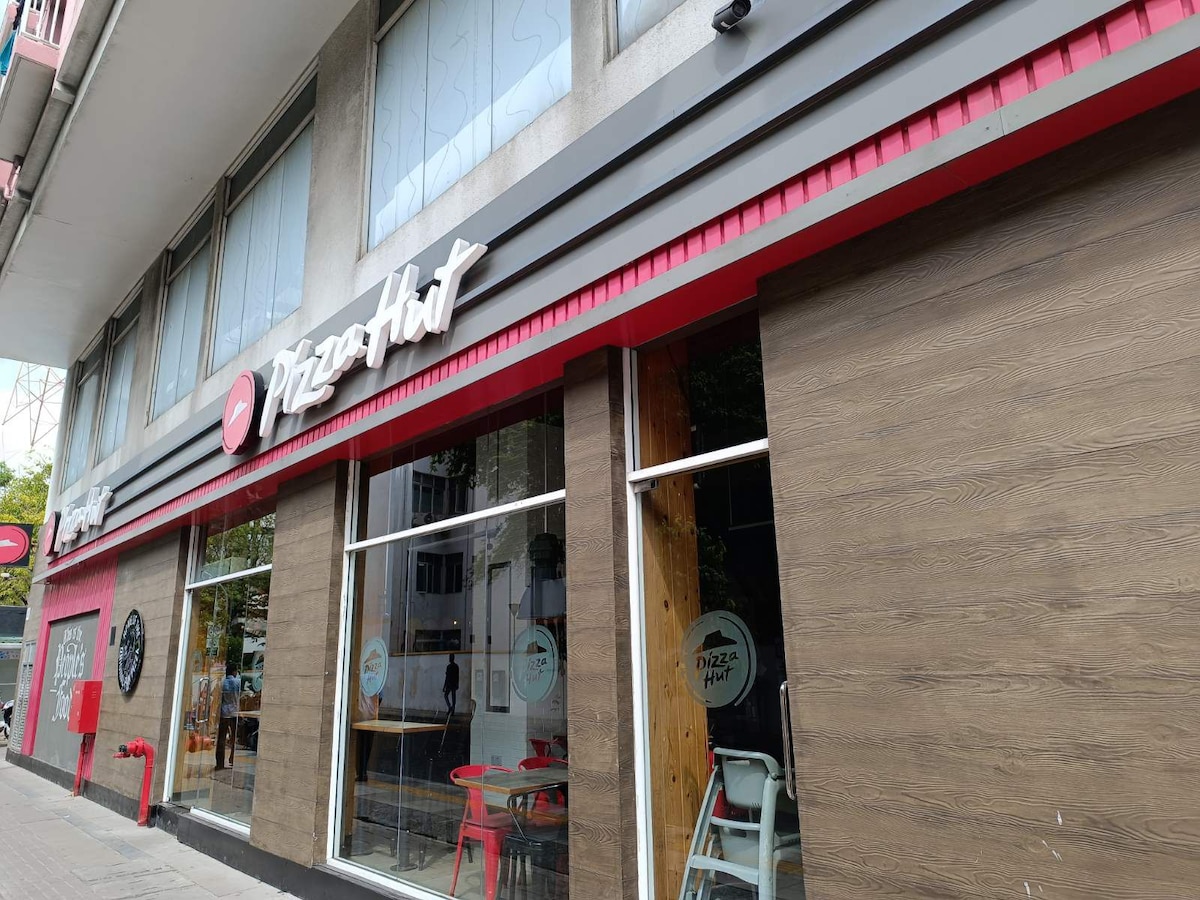 A modern restaurant exterior is displayed, featuring large glass windows with a red and white sign displaying the name 'Pizza Hut.' Wooden accents complement the contemporary design, while outdoor seating is visible in front, inviting guests to enjoy the vibrant street view.