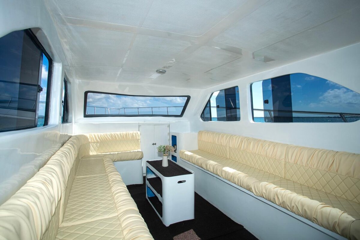 The interior of the yacht is furnished with soft, cream-colored seating arranged around a central table. Large windows provide ample natural light and views of the ocean, creating an open and airy atmosphere.