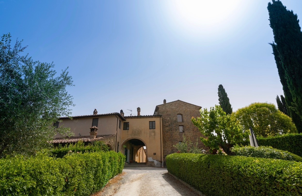 The entrance to the property features an arched gateway flanked by well-manicured hedges and trees. A gravel path leads towards the main building, which displays a rustic stone façade. The sky above is clear, creating a bright and airy atmosphere.