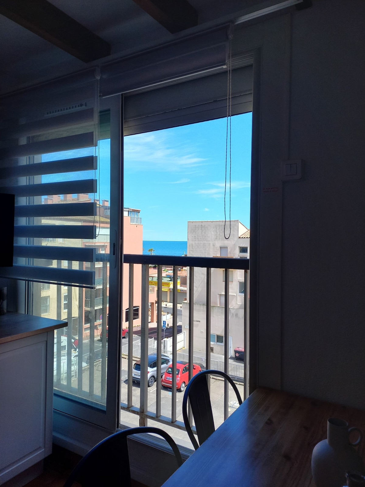 A view from the apartment reveals a clear blue sky over the ocean. The glass door features modern blinds, and a wooden dining table with black chairs is positioned near the window, allowing natural light to fill the space.