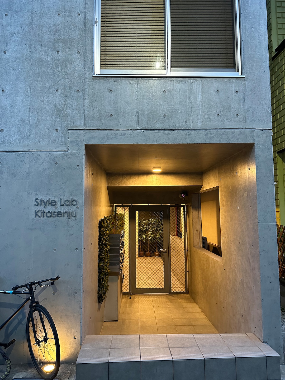 The entrance to the building is characterized by a sleek concrete facade, featuring large glass doors behind a protective metal gate. A bicycle is parked nearby, and soft lighting illuminates the area, creating a welcoming atmosphere.