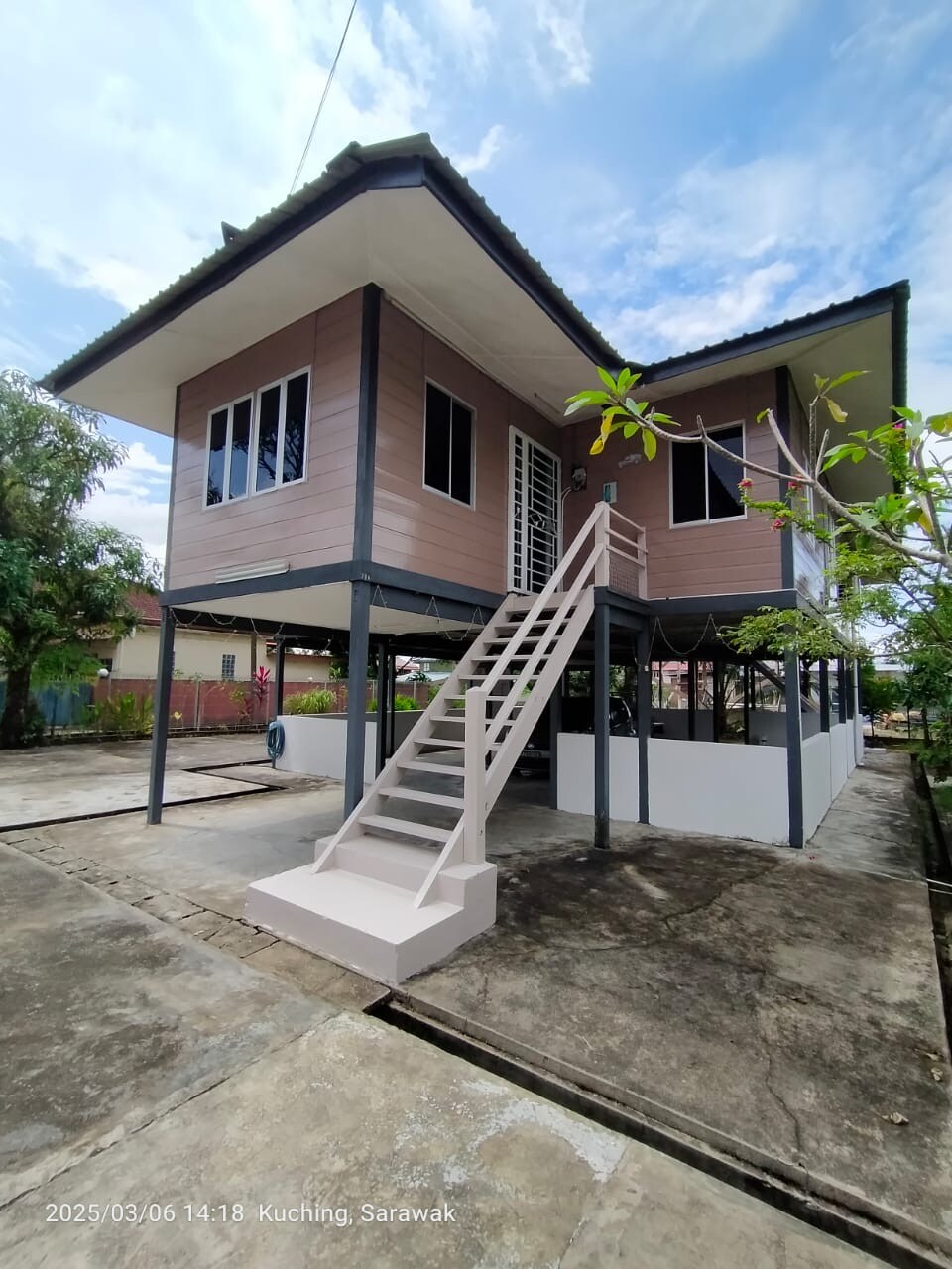 A two-story house is situated on a concrete foundation, elevated above ground. A set of stairs leads to the main entrance, which is framed by large windows. Surrounding greenery and an open area are visible, contributing to a spacious outdoor environment.