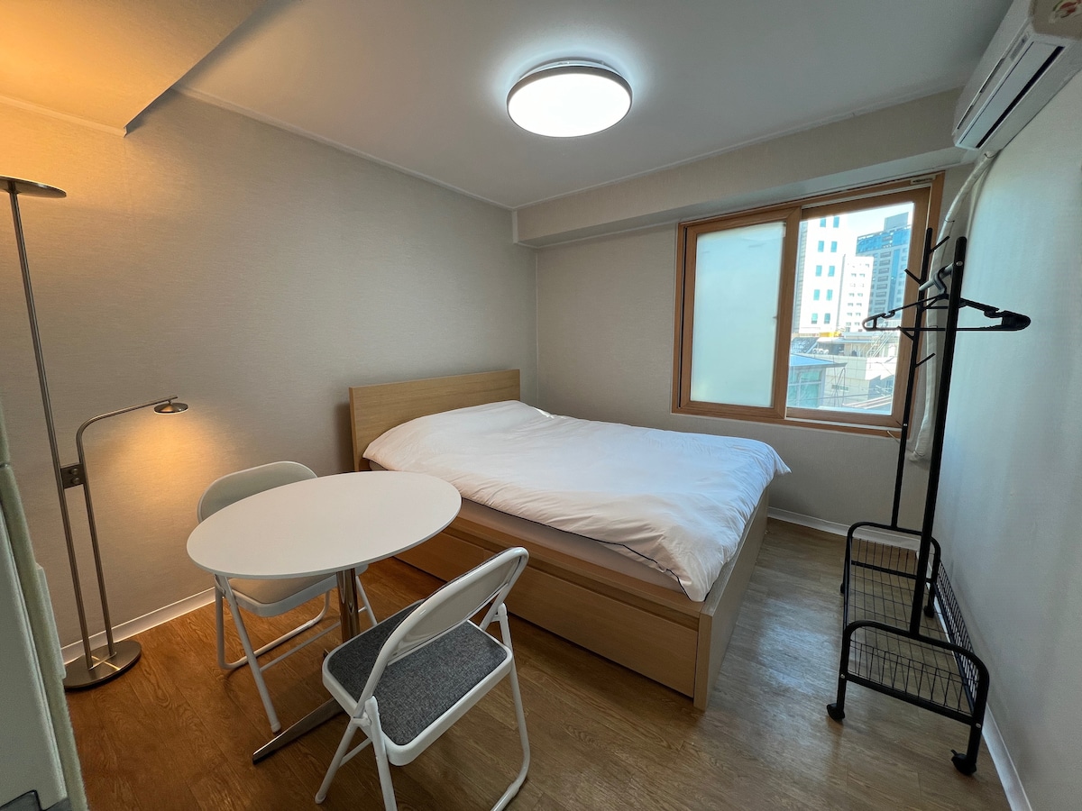 The room features a spacious bed, a round table with two chairs, and a window allowing natural light to fill the space. A clothing rack is positioned nearby, and an air conditioning unit is installed above the bed, providing comfort to the area.