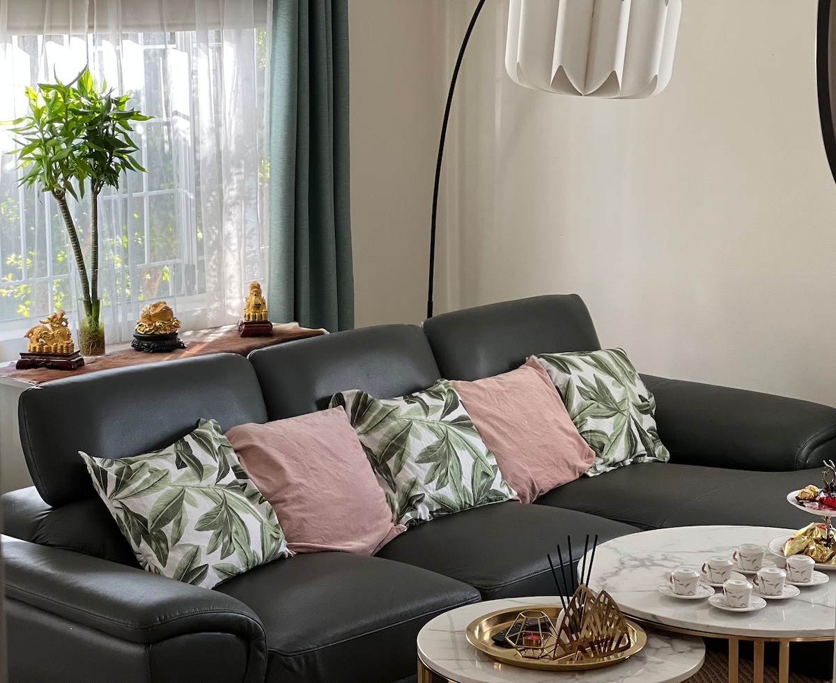 A comfortable leather sofa is adorned with cushions featuring leafy patterns and soft pink tones. A marble coffee table holds tea sets and decorative items. Natural light streams in through a window, complemented by green curtains, and a potted plant is positioned nearby.