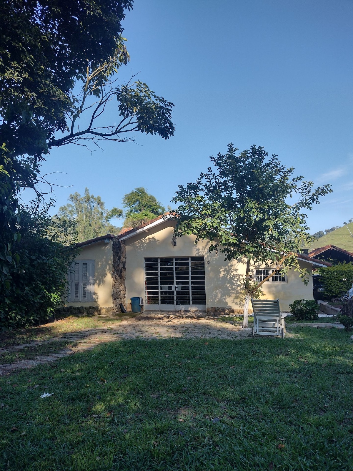 The exterior of a charming single-story house is framed by lush greenery. A tree stands beside the house, while a stone path leads to a seating area on the well-maintained lawn. Bright sunlight bathes the scene, contributing to a peaceful environment.