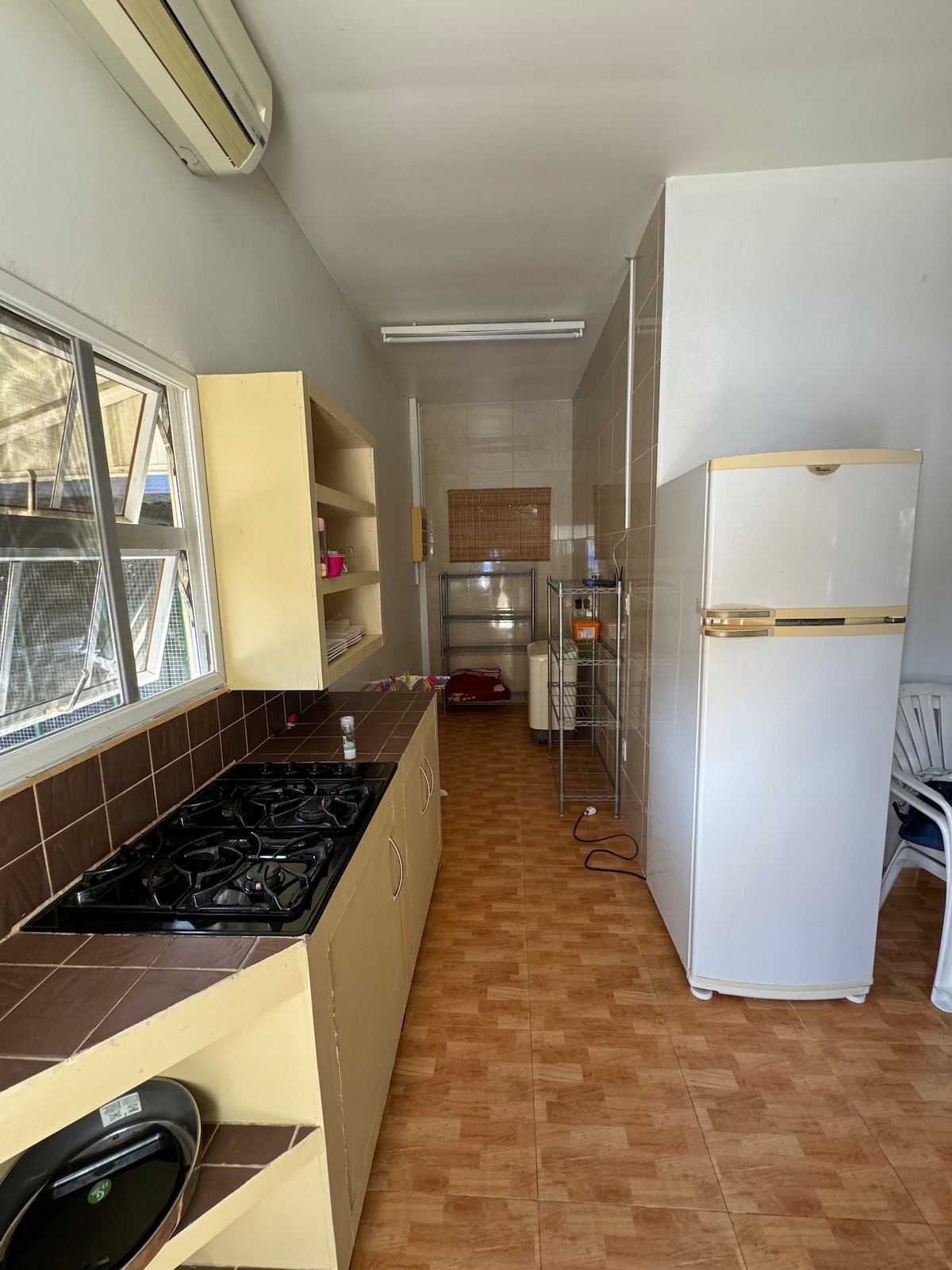 A compact kitchen area is visible, featuring beige cabinetry and a dark countertop with a four-burner stove. The space is illuminated by natural light through a window. A white refrigerator stands against the wall, and a few culinary items are placed along the countertop.
