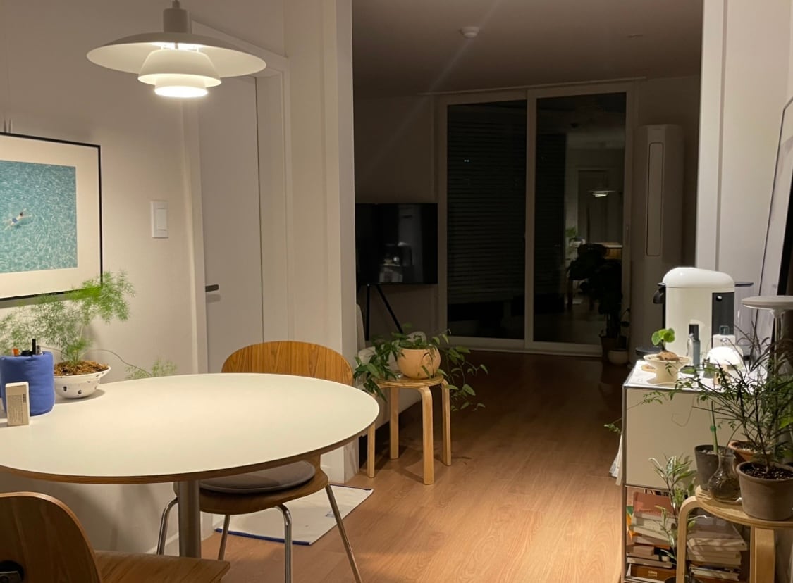A cozy dining area is fitted with a round table and two wooden chairs, surrounded by plants. A framed artwork is displayed on the wall, while soft lighting casts a warm glow. In the background, large glass doors reveal a well-lit living space.