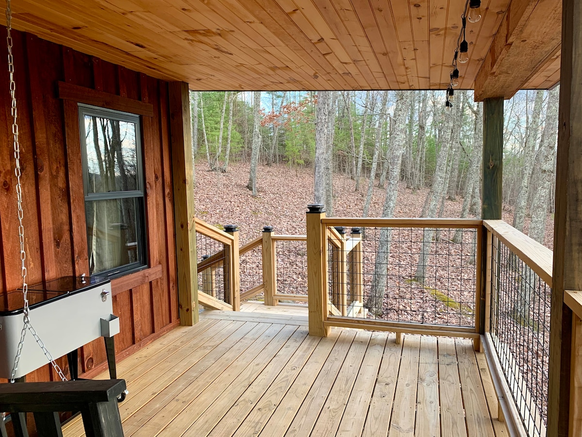 A spacious back deck features wooden flooring and railings with a natural finish, surrounded by tranquil wooded scenery. Seasonal leaves carpet the ground below, and string lights hang overhead, creating a peaceful space for relaxation.