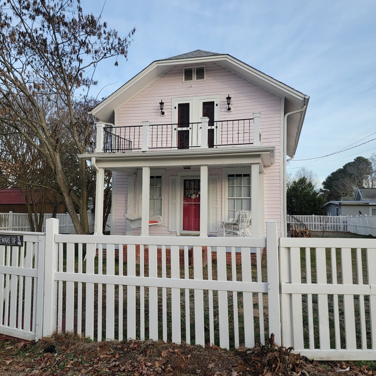 A charming two-story house features a light pink exterior, complemented by white railings and a welcoming red front door. A white picket fence surrounds the yard, while a few trees and shrubs add a touch of greenery. Two rocking chairs are positioned on the front porch.