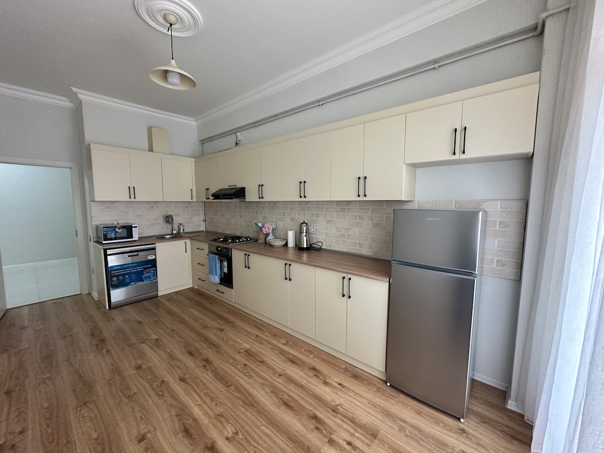 A modern kitchen features light-colored cabinetry and a spacious countertop. Stainless steel appliances, including a refrigerator and stove, are visible. Natural light filters through sheer curtains, reflecting off the smooth laminate flooring.