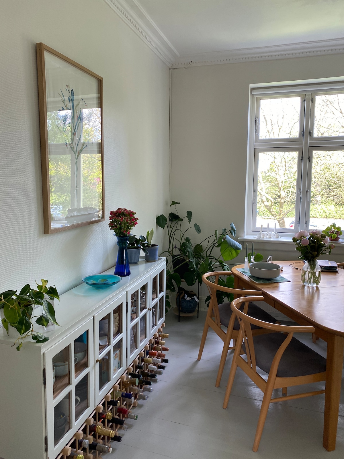A dining area features a wooden table surrounded by light chairs. A white console with glass doors displays an assortment of plants and decorative items. Natural light enters through a large window, enhancing the greenery and floral arrangements throughout the space.