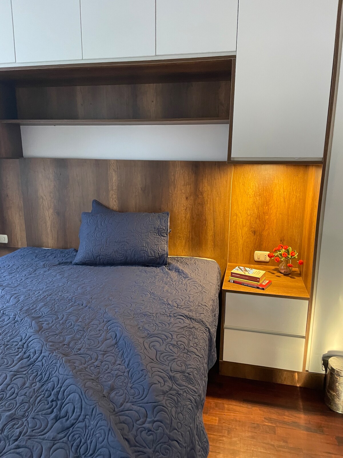 A cozy sleeping area features a spacious bed topped with a dark blue quilted cover. A wooden headboard and bedside table are visible, with a small vase of flowers and a few books on the table. Soft lighting highlights the wooden surfaces.