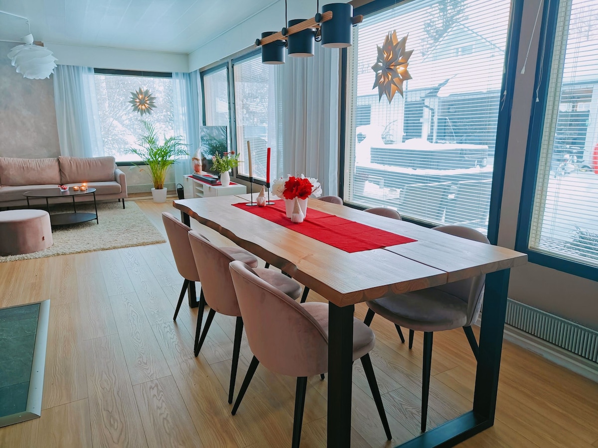 A modern dining area features a large wooden table surrounded by upholstered chairs. A red table runner with a floral centerpiece is visible, complemented by decorative candles. Expansive windows allow natural light to fill the space, showcasing views of the outside while providing a cozy atmosphere.