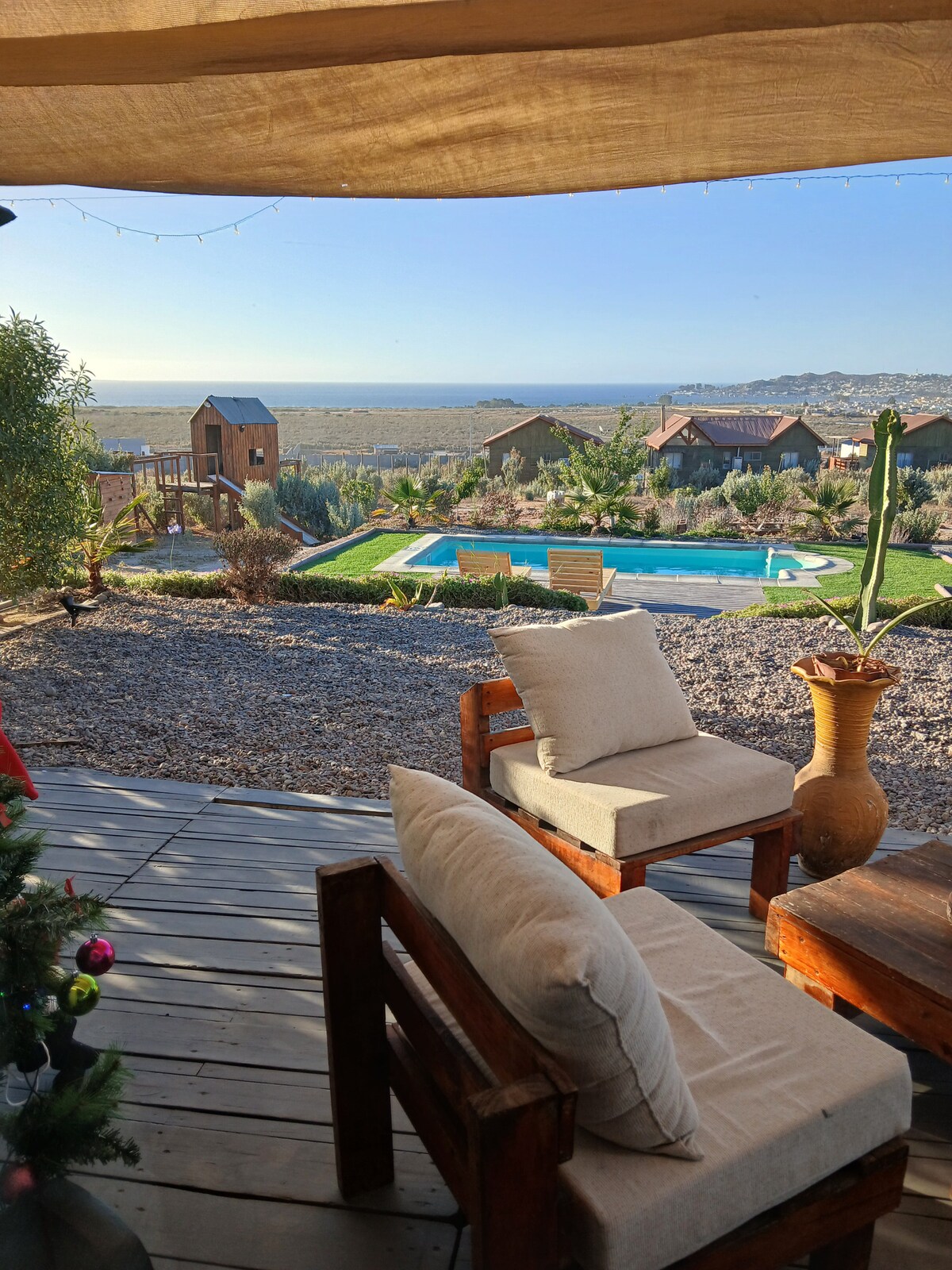 A shaded outdoor seating area features two cushioned wooden chairs, with a view of a serene swimming pool surrounded by landscaped gardens. The backdrop includes a play area and distant cabins, while the landscape extends towards the horizon, providing a peaceful, natural setting.
