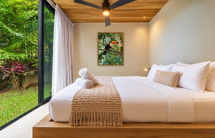 Pangunahing kuwarto sa lower suite! Isang tahimik na silid-tulugan na may queen-size na higaan at mga premium na linen na may kalidad ng hotel, mga handcrafted na muwebles na gawa sa kahoy, orihinal na tropikal na likhang-sining ni Tylaya, at magagandang tanawin ng hardin—na lumilikha ng mainit at nakakarelaks na kanlungan na napapaligiran ng kalikasan