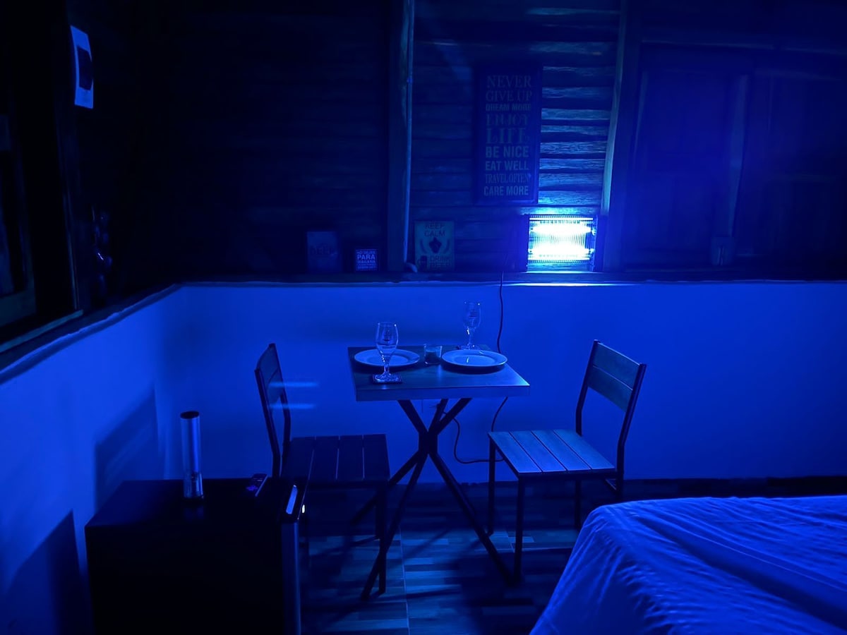 A small dining area features a table set for two with glasses and plates. Two wooden chairs are positioned around the table, while a sleek fridge stands nearby. Soft blue lighting creates a calming ambiance within the room, highlighting the wooden wall elements and window frames.