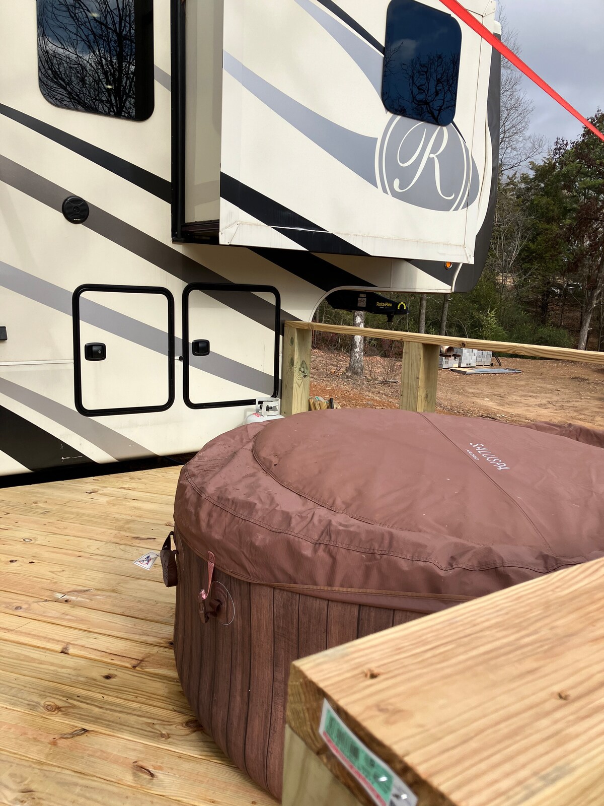 A spacious deck is featured, showcasing a hot tub with a brown cover. The area is framed by the exterior of the property, which displays sleek lines and a large awning. Surrounding trees add to the peaceful outdoor setting.