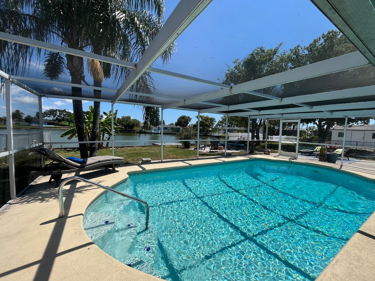 A private pool is surrounded by a screened enclosure, with clear blue water reflecting sunlight. Lounge chairs are positioned nearby, and palm trees are visible in the background, enhancing the serene lakeside view.