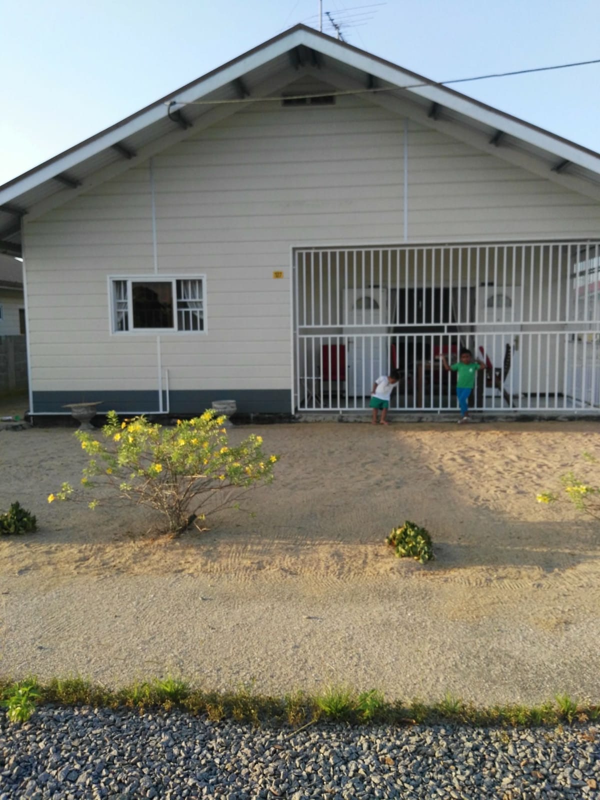 A single-story house is depicted, featuring a welcoming facade with security bars across the front entrance. Two children can be seen engaging playfully in the yard, which has simple landscaping and gravel paths. The neighborhood appears quiet and the house maintains a neutral color scheme.