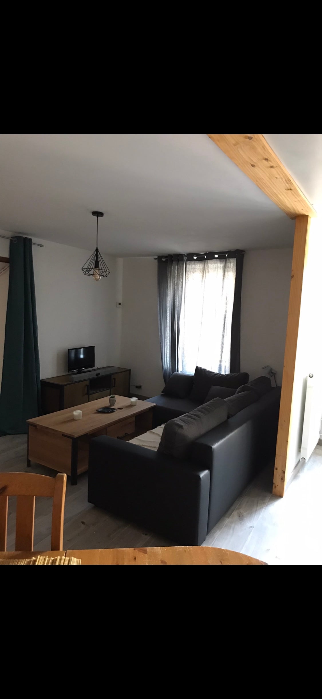 A cozy living area features a black leather sofa with gray cushions, positioned near a wooden coffee table. A small TV is placed on a media console. Light filters through large windows, accented by light curtains, creating a bright and inviting space.