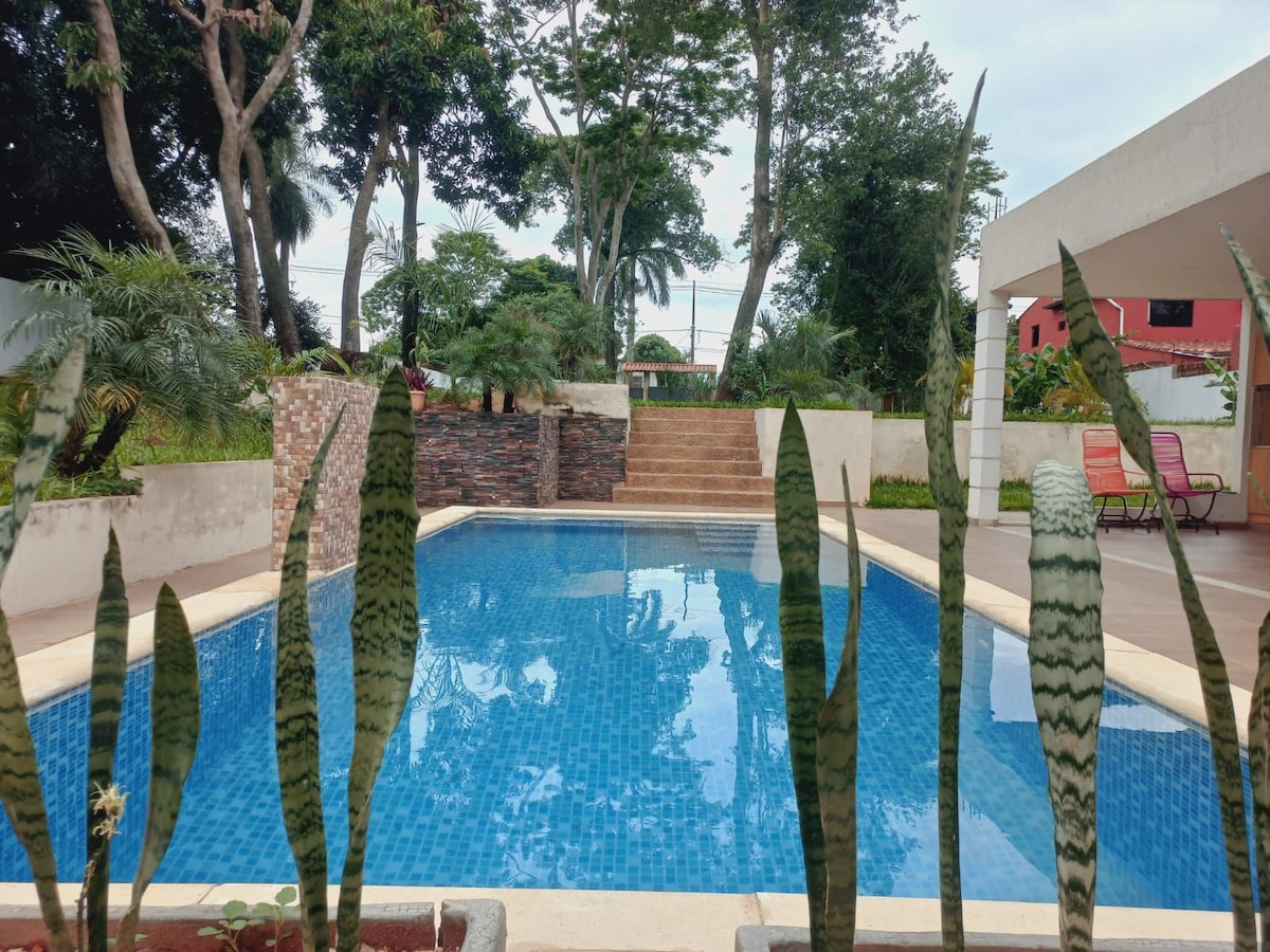 A clear swimming pool is framed by green plants and trees, providing a serene outdoor space. Steps lead up from the pool area to a landscaped section. The atmosphere is peaceful, inviting relaxation under the open sky.