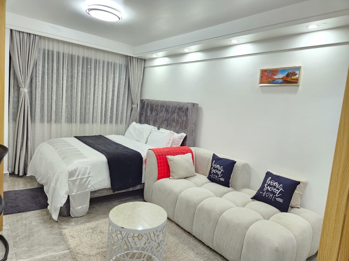 A modern studio features a spacious bed with crisp white bedding and a decorative throw. A light-colored sofa, adorned with cushions, complements a round coffee table. Natural light filters through sheer curtains, enhancing the welcoming ambiance of the room.
