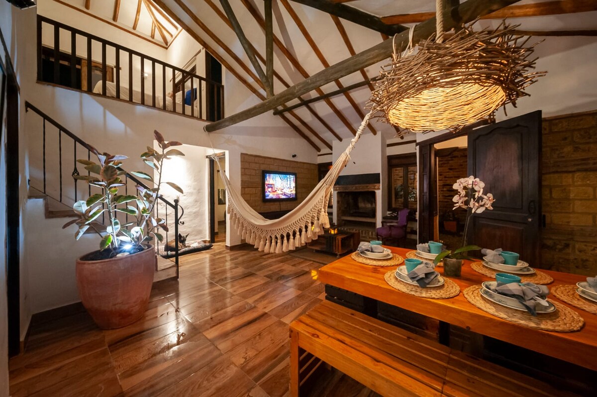 The living area features a high ceiling supported by wooden beams, complemented by a cozy hammock and a rustic dining table set for six. A television is mounted on the wall, and a fireplace adds warmth to the space. Natural light filters through from the windows.