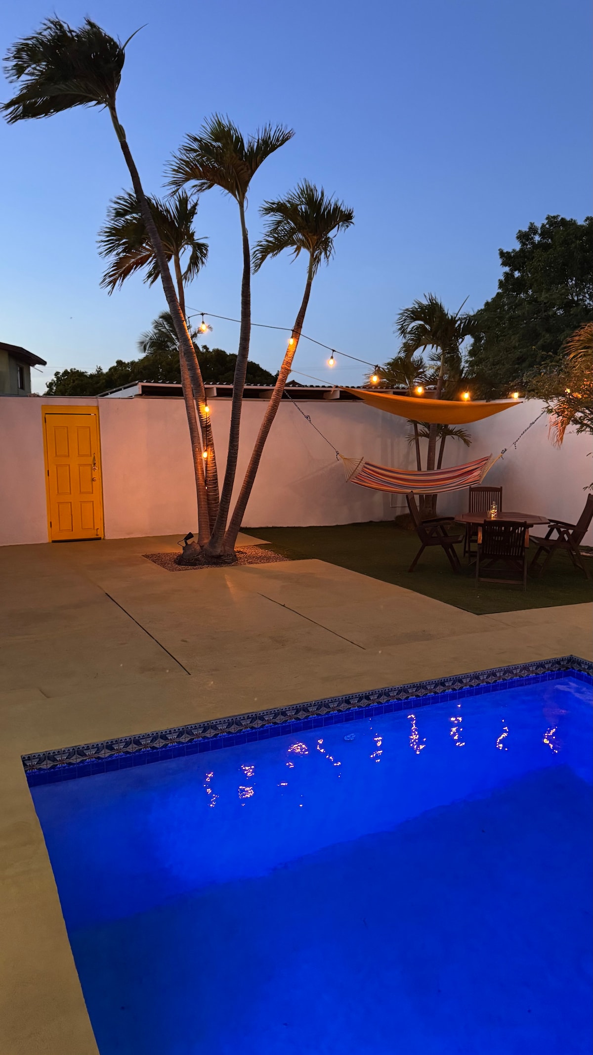 A private outdoor area features a sparkling pool with illuminating water reflecting the evening sky. Palm trees sway gently nearby, and soft string lights create a serene ambiance. Nearby, a hammock and a dining set invite relaxation and social gatherings under the stars.