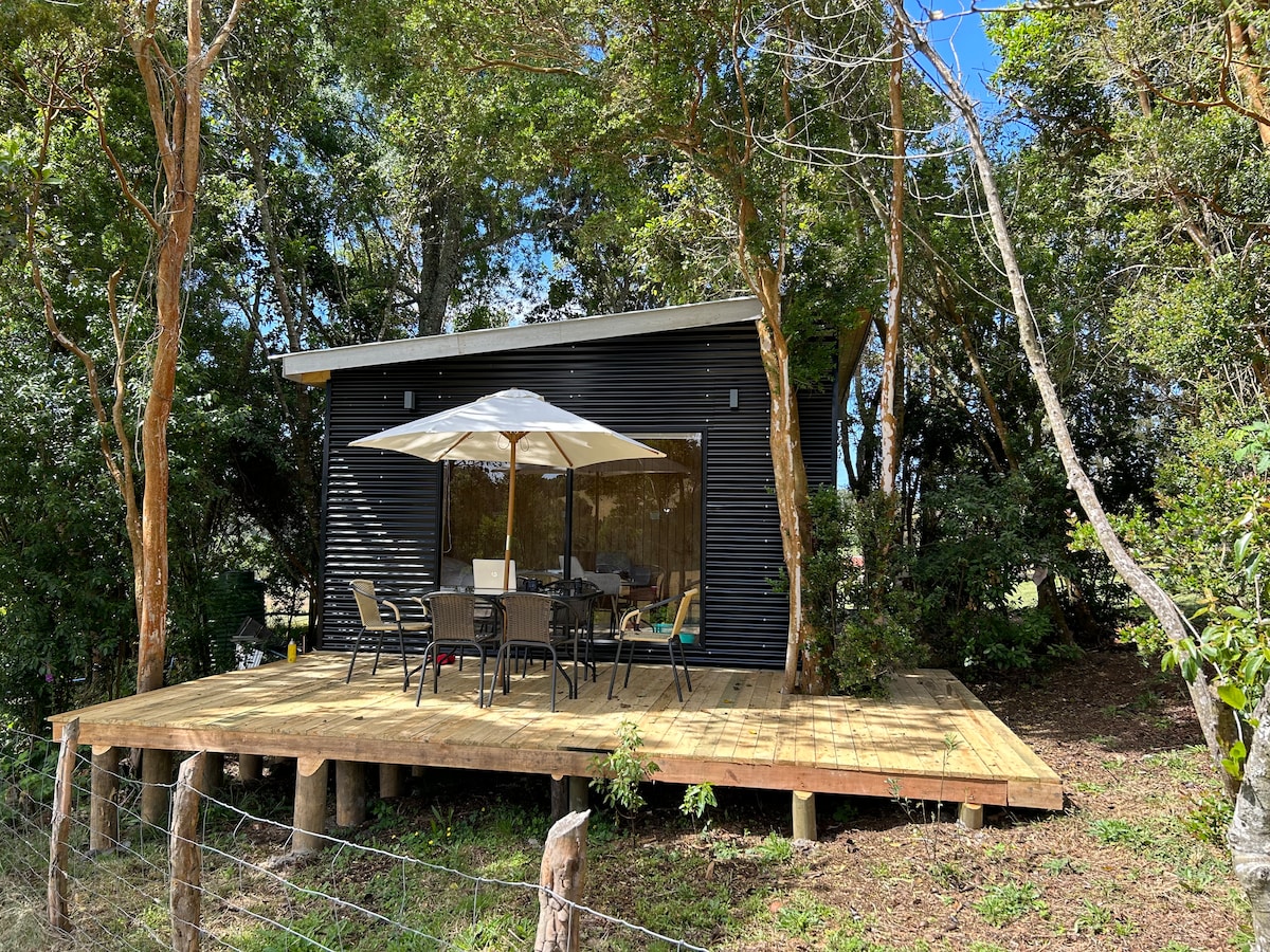 A modern cabin is surrounded by native trees, featuring a spacious wooden deck. A large umbrella provides shade over a dining table with several chairs, offering an inviting space for outdoor relaxation in a natural setting.