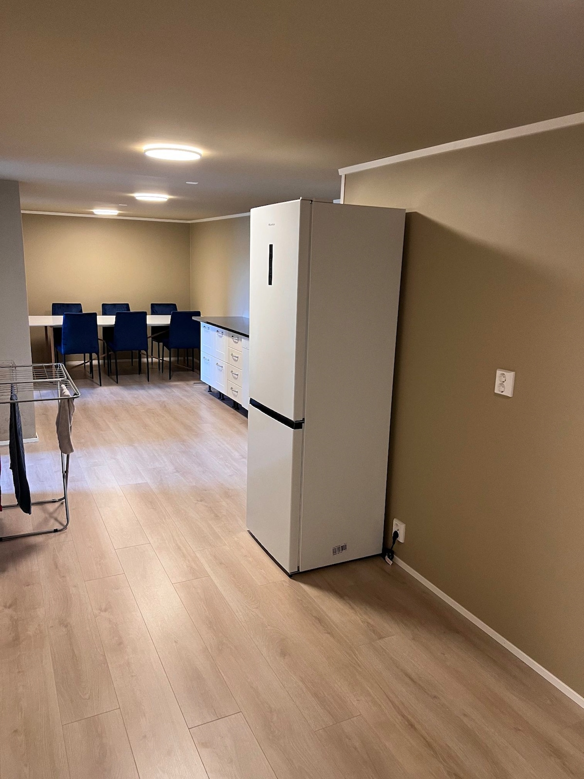 A modern kitchen area is featured, showcasing a tall white refrigerator positioned against a neutral-toned wall. A dining table with blue chairs is visible in the background, complemented by sleek cabinetry. Natural light enhances the warm wooden flooring throughout the space.