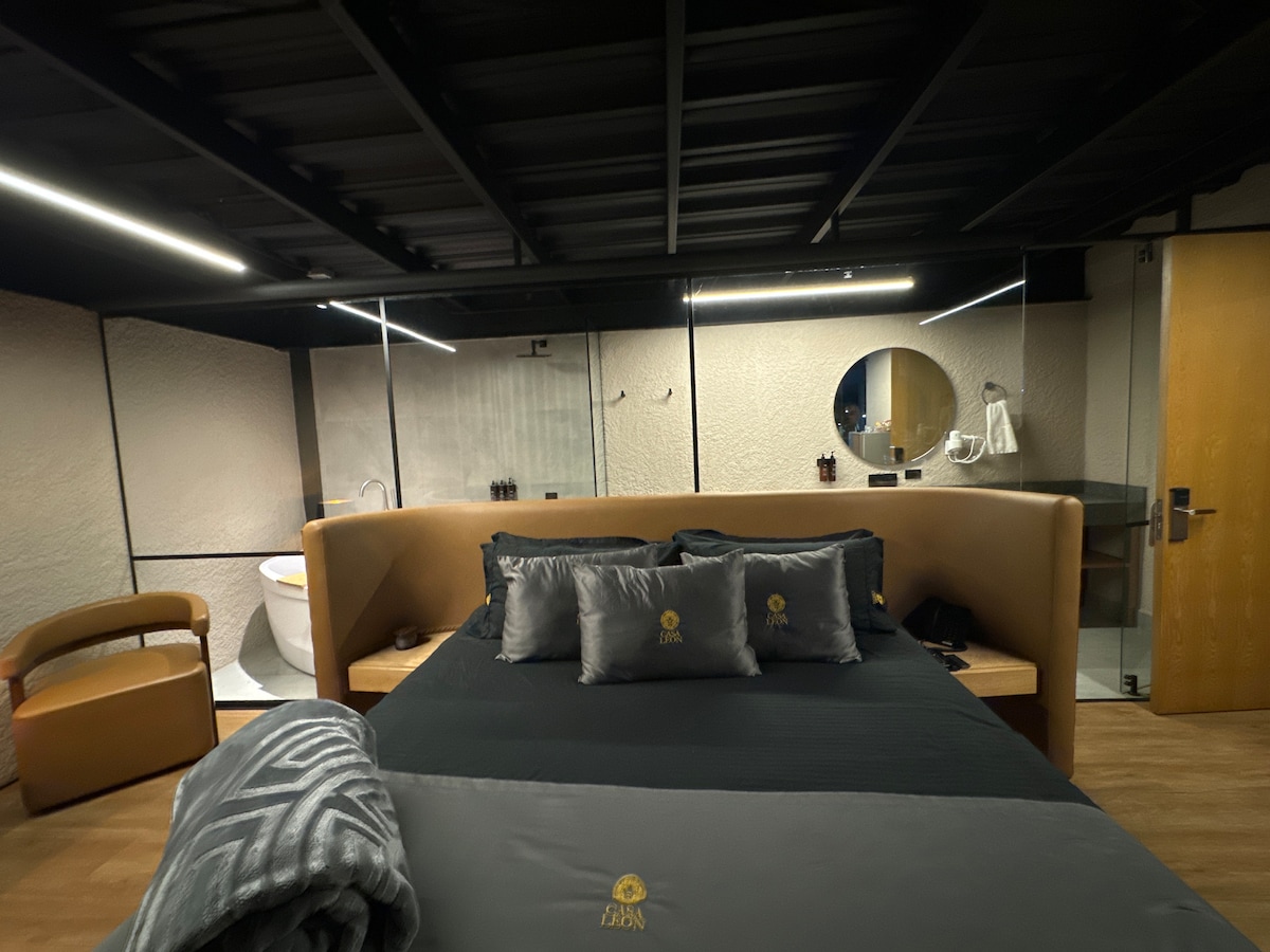 A stylish suite features a spacious bed adorned with decorative pillows and a soft throw. The room is complemented by a modern bathroom area visible in the background, as well as a freestanding tub and minimalist furnishings, providing a contemporary aesthetic.