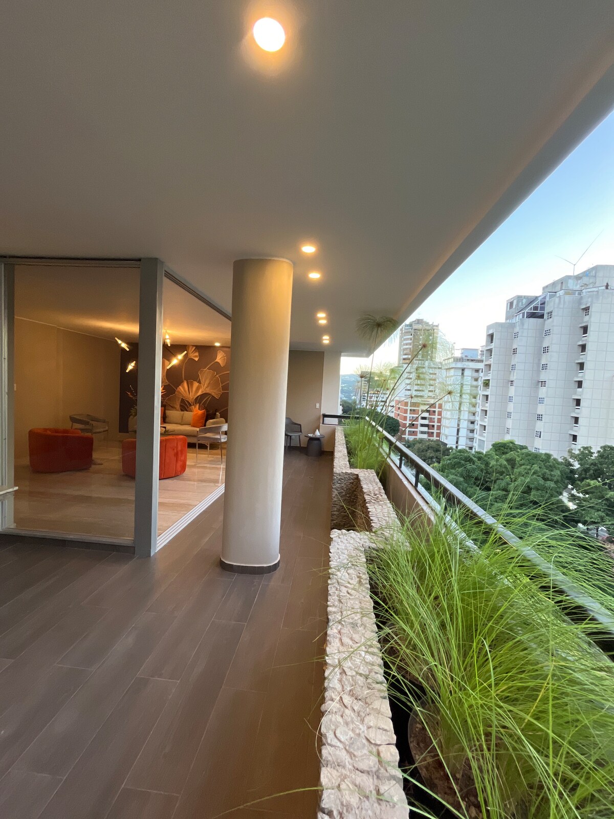 A spacious balcony features a seamless connection to the indoor living area. Potted plants line the edge, contributing to a natural ambiance. The view includes neighboring buildings and greenery, with soft lighting enhancing the overall setting.