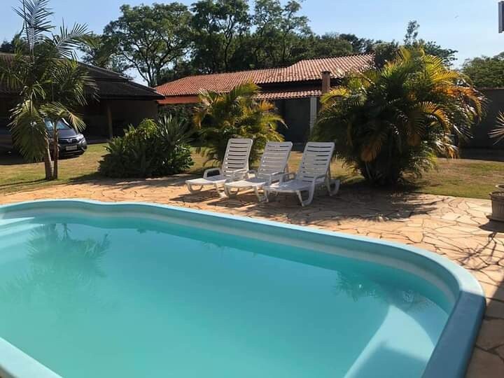 A serene outdoor space features a clear pool bordered by stone flooring. White lounge chairs are arranged nearby, offering a comfortable spot for relaxation. Lush green plants and palm trees add to the natural surroundings, creating a peaceful environment for family gatherings.