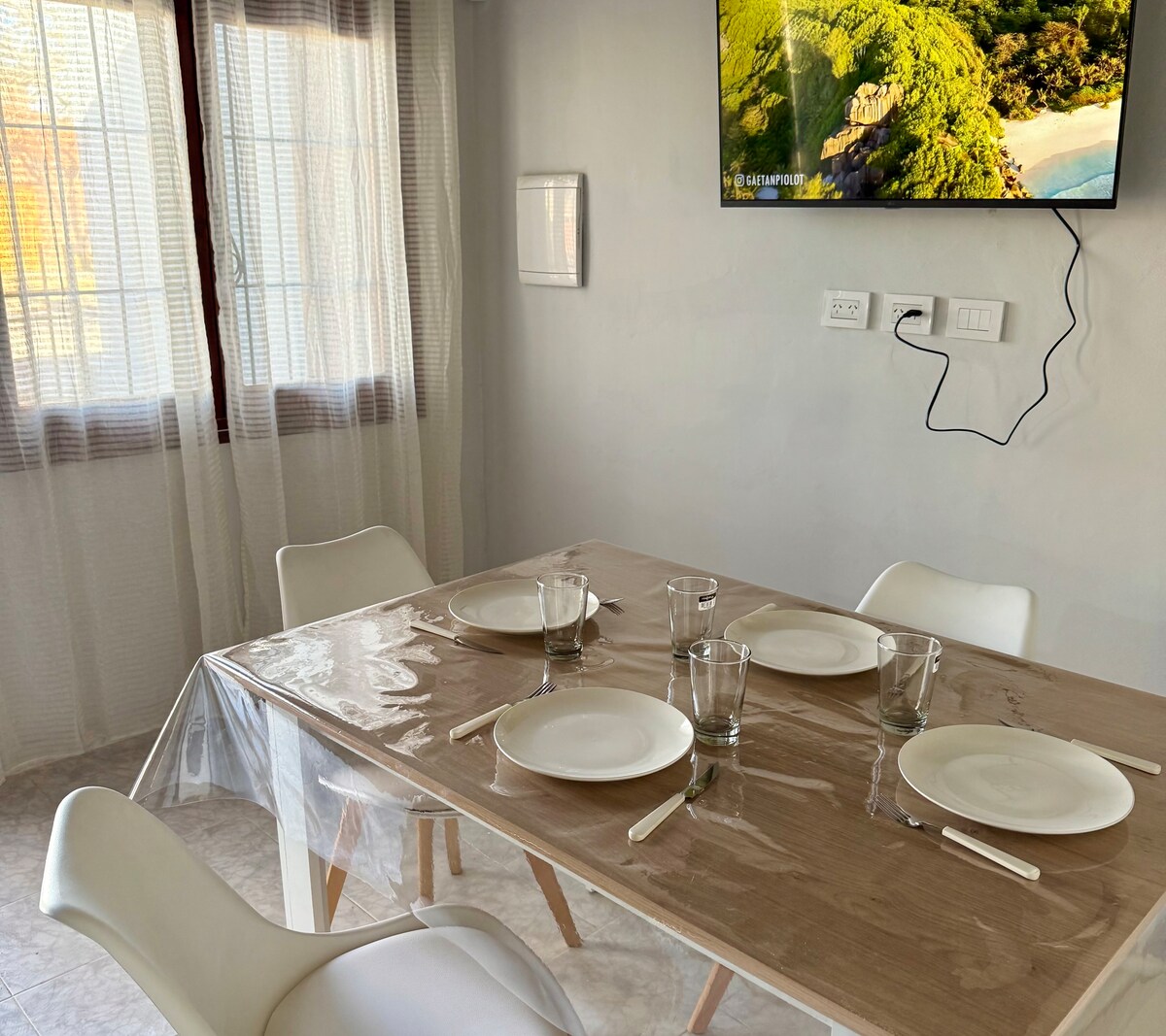 A dining area features a wooden table with a clear protective covering. Four white plates and matching glasses are placed at each setting. Light drapes frame the window, while a television displays a serene natural landscape on the wall.