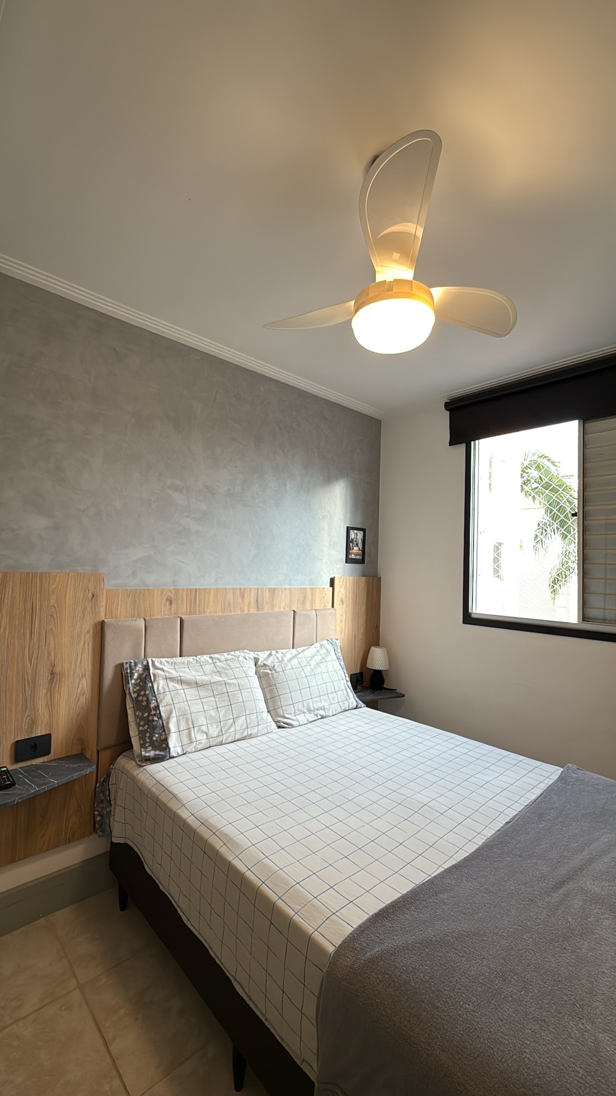 A cozy bedroom features a spacious bed with light linens and a soft throw blanket. A ceiling fan provides airflow, while a window lets in natural light, partially obscured by a sleek window treatment. Nightstands flank the bed, enhancing the organized space.