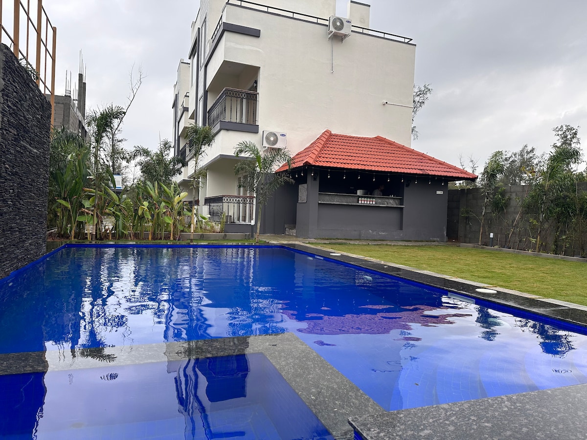 A serene swimming pool is surrounded by lush greenery and a well-maintained lawn, featuring a dark stone border. In the background, a two-story building with a red-tiled roof and outdoor bar area adds to the tranquil atmosphere.