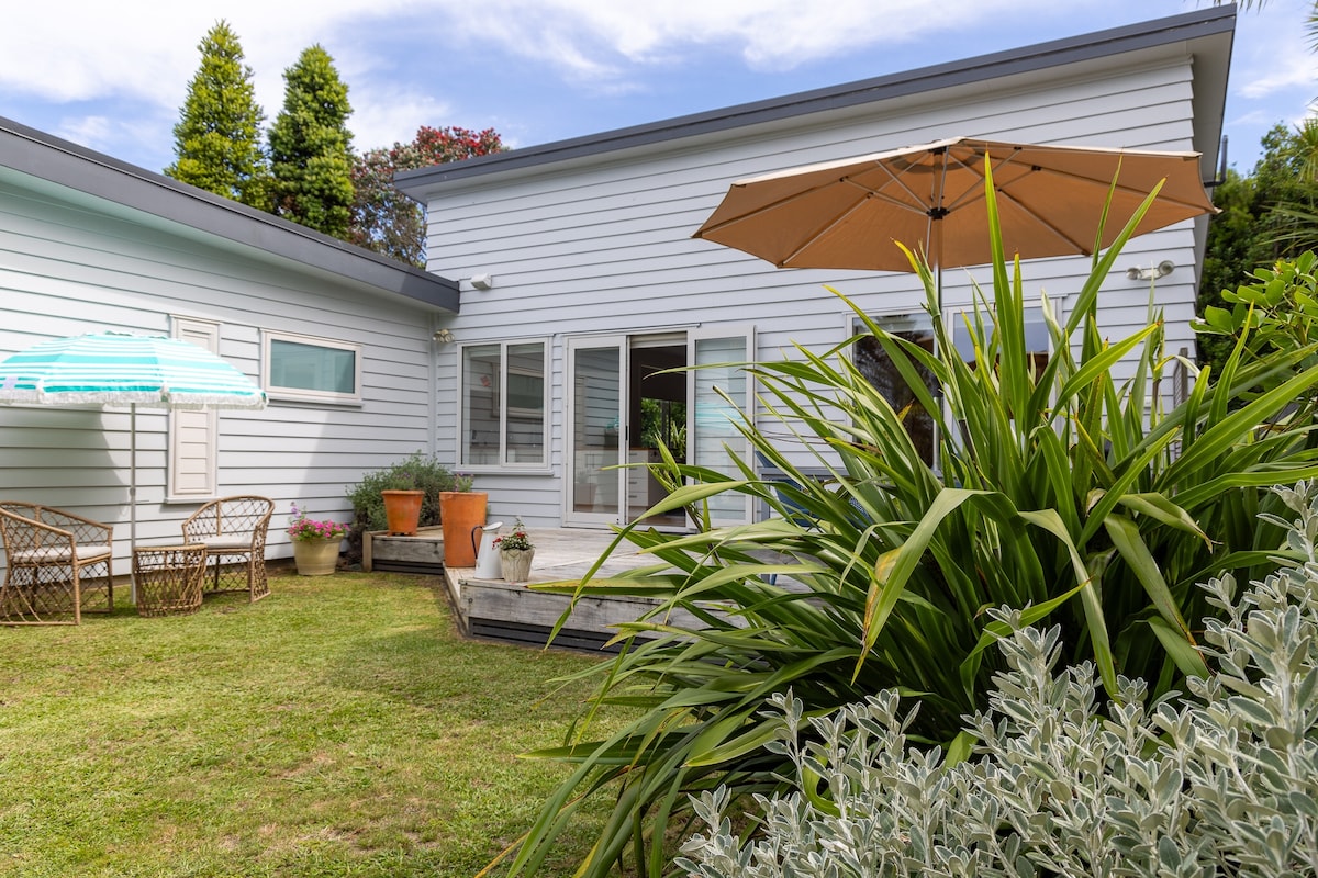The exterior of the guest house is seen with a sunny outdoor area, featuring a patio space and an umbrella. Lush greenery surrounds the space, including potted plants and well-maintained grass, providing a serene atmosphere for relaxation.
